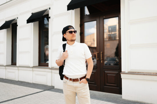 Caucasian Man With Backpack Walking Through Historic Streets, Casual White Tshirt And Cap, Backpack Slung