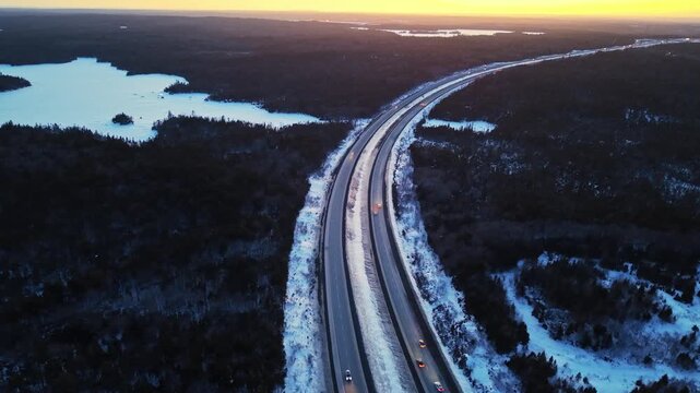 High-angle drone view: vehicles with bright headlights move continuously along a snowy highway during blue hour. Winter traffic flow in Halifax region, Nova Scotia, Canada 4K.