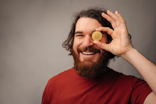 Bearded Man Holding Gold Bitcoin Coin, Grinning At Camera In Casual Red Shirt, Curly Hair And Warm Studio