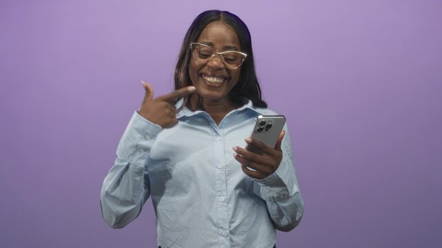 Woman points finger to chest while holding smartphone in studio purple backdrop wearing glasses and light blue shirt, smiling broadly; confidence.