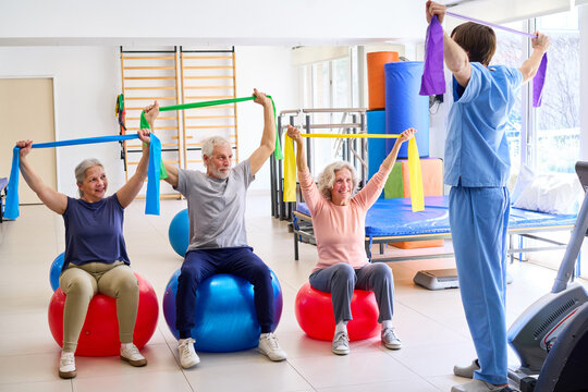 Senior Adults Doing Exercises With Resistance Bands On Exercise Balls In Rehab Center With Physiotherapist Supervising For Rehabilitation