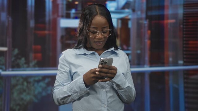 Black woman wearing glasses holds a smartphone with both hands, tapping and reading messages while standing near glass railing in a modern building; focus connection.