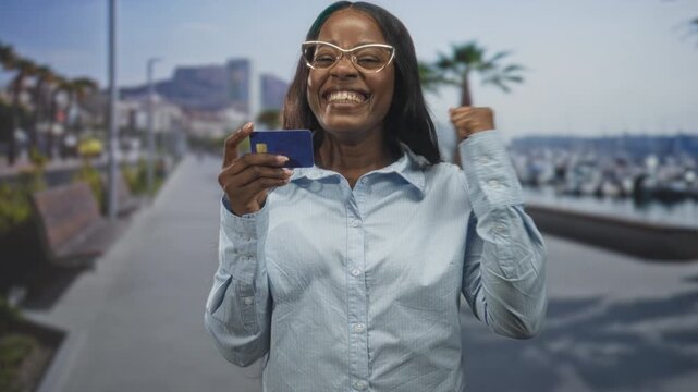 Woman holds creditcard with raised fist on street pier near marina, smiling and celebrating a successful payment; celebration success.