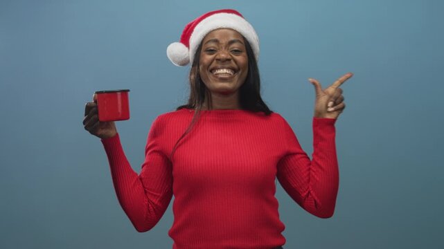 Woman wearing santa hat smiles broadly, holds red mug and points finger to side in studio; holiday joy.