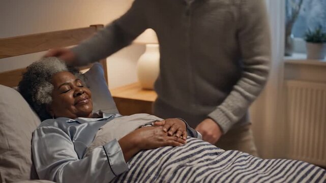 Caregiver tucks a smiling senior African American woman into bed at home.