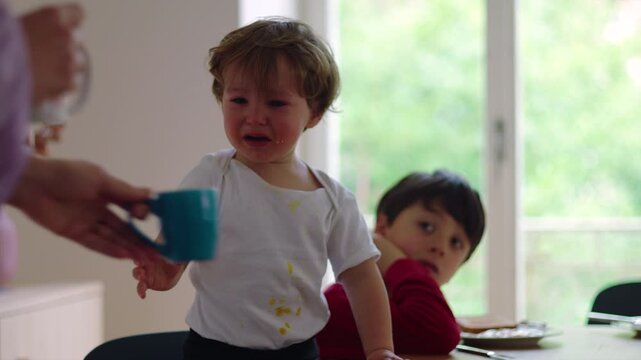 Young child standing at table crying and pointing forward during mealtime while older sibling sits behind appearing quiet and withdrawn in home environment