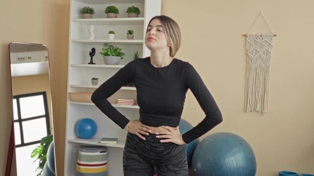 Young woman hands on hips showing bare waist and stretching torso in studio with exercise ball, mirror and shelving; serenity.