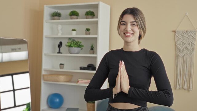 Young woman smiling with palms pressed together in prayer pose wearing black crop top in a home yoga studio with plants and mirror; serenity.