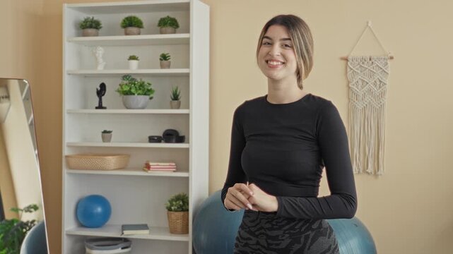 Woman with exercise ball smiles with clasped hands in a home studio beside shelving and macrame wall hanging; wellness serenity.