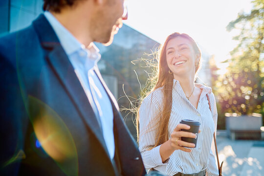 Two smiling business colleagues having a casual outdoor conversation, enjoying a coffee break in bright sunlight