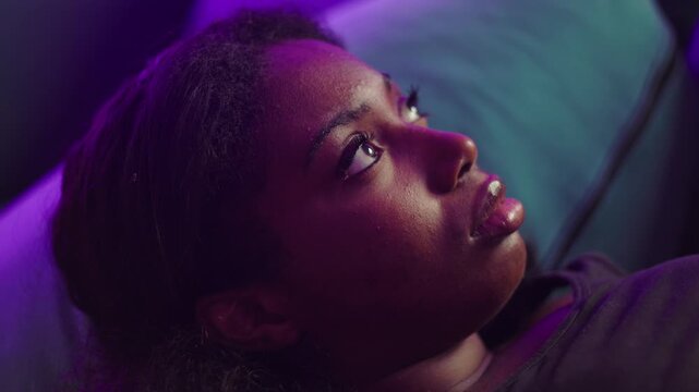 African American woman lying on couch with distant gaze reflecting on past mistakes feeling regret and sadness while lost in quiet introspective thought indoors