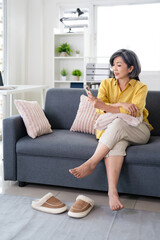 Obraz premium Happy middle aged asian woman using smartphone sitting on couch at home. holding cellphone browsing internet, texting messages on mobile cell phone technology.
