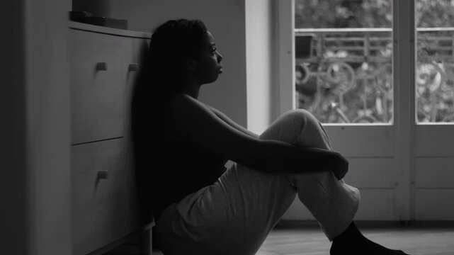 African American woman sitting on floor hugging knees by window in monochrome scene conveying deep depression vulnerability isolation and psychological distress at home