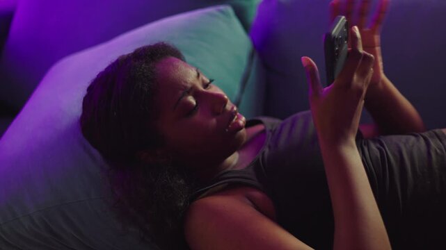 African American woman reclining on sofa engaging with smartphone before shifting into disapproval, distancing from device as reaction to unwanted message or content in quiet home environment