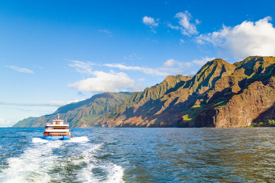 A motion blurred tour boat cruises through the deep blue waters of the Pacific, leaving a white wake as it passes the dramatic, emerald-green fluted cliffs of Kauai's stunning Na Pali Coast.