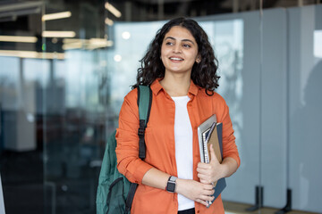 Young indian woman carrying a backpack, laptop, and notebooks, smiling and looking away while...