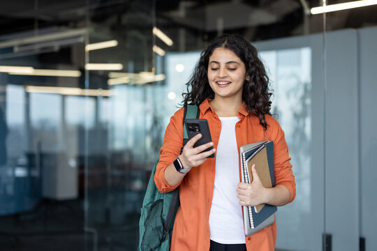 Young indian female student carrying a backpack and notebooks, smiling while looking at her mobile phone, walking through a modern university building