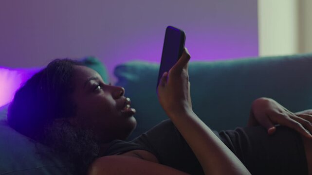 African American woman lying on couch speaking into smartphone while sending voice message, relaxed posture and casual communication during quiet evening at home