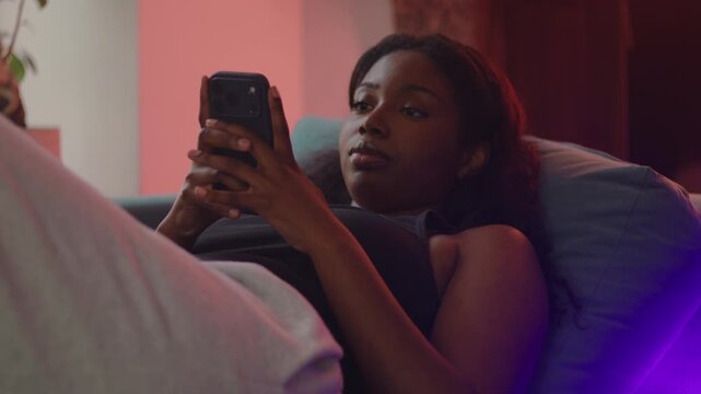 African American woman lying on couch using smartphone as expression shifts from neutral to smile, reacting to message or content, capturing emotional change and positive digital interaction at home
