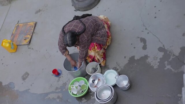 A cholita, a woman in a wide, long skirt, rinsing kitchen utensils in a courtyard.