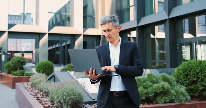 Confident businessman using a laptop outdoors near a modern office building, working remotely and managing online business.
