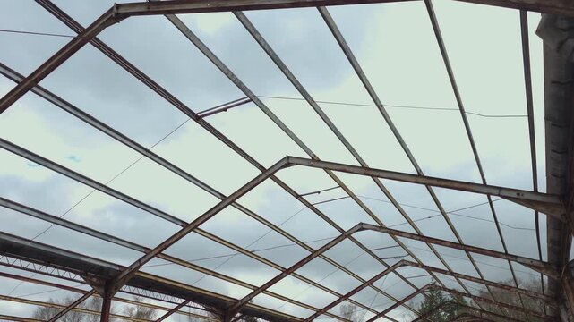 Derelict airbase hangar interior with skeletal metal construction and cloudy sky above. Drone perspective moving beneath corroded beams tangled wires and aging steel structure in forgotten military