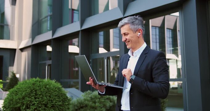 Portrait of a focused businessman in a formal suit conducting an online conference on a wireless laptop outdoors.