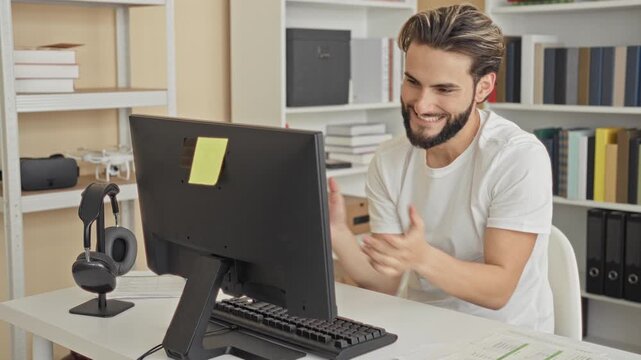 Young man at computer smiling and pumping fists at desk with keyboard and monitor in a studio office with bookshelves and storage boxes behind him; satisfaction focus.