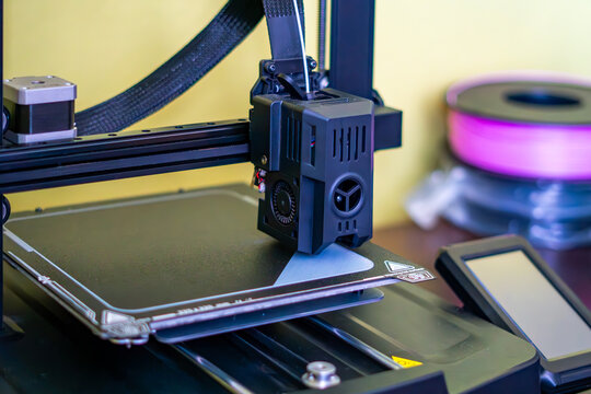 3D printer nozzle printing first layer of a model on a magnetic bed. FDM technology in action, additive manufacturing process, pink filament spool in background. Prototyping, engineering and modern te