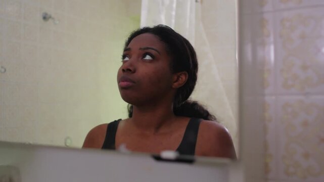 African American woman looking down at mirror with troubled expression, inner doubt and emotional struggle during quiet bathroom moment, reflecting insecurity and personal tension