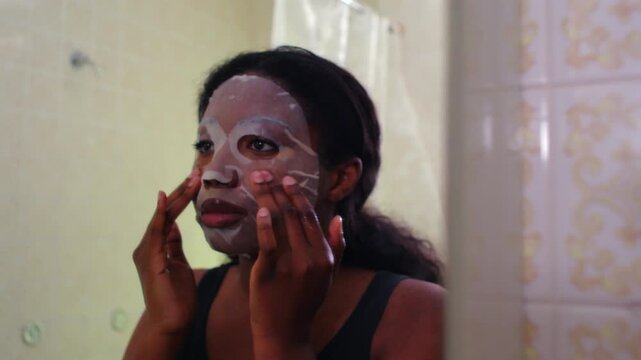 African American woman adjusting facial sheet mask with both hands in bathroom, centered view of skincare routine, calm focused expression and careful placement of beauty treatment