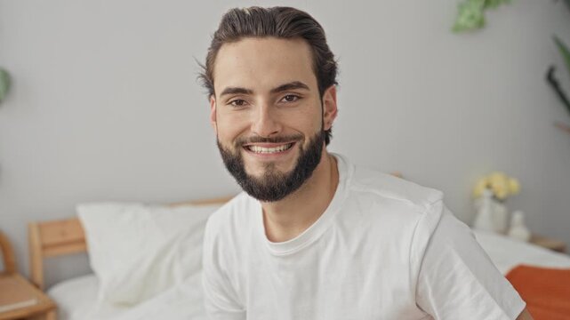 Man sitting on bed smiling, face and beard visible, wearing white tshirt in a studio bedroom setting with plants, bedside table and bedding; serenity morning.