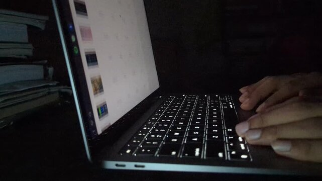 Person typing on illuminated keyboard of laptop computer in dark room, scrolling through data and charts showing business growth and sales performance.