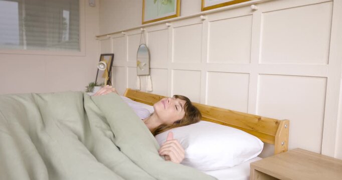 Happy mature woman waking up in the morning, snuggling comfortably under a warm duvet
