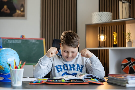 Young boy celebrating an achievement while reading an open book at his study desk and learning in a home environment