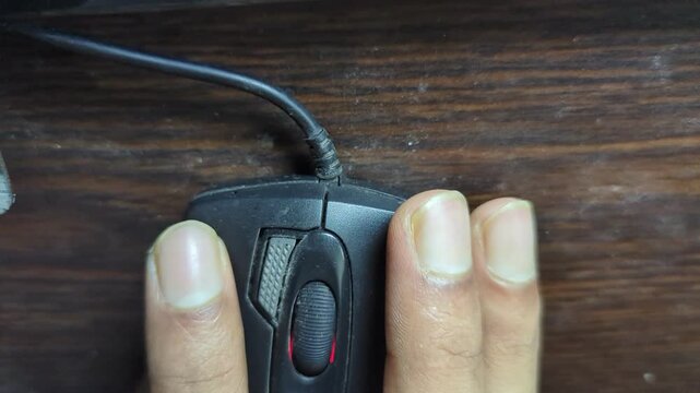 Close-up of a human hand scrolling and clicking on a black computer mouse on a wooden table, with the red light of the mouse visible and fingers in motion during computer operation.