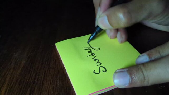 Person writing sunday with black marker on yellow sticky note paper placed on dark wooden desk surface close up of hand holding felt tip pen creating handwritten text in natural light indoors.
