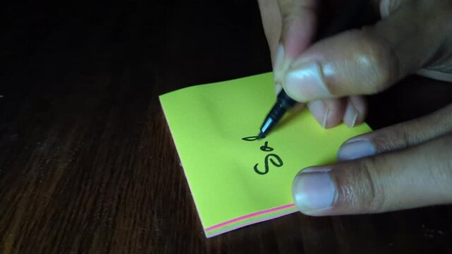 Person writing saturday on yellow sticky note with black marker on wooden desk surface close up of hand holding pen and lettering day of week in cursive style for planning and organization.