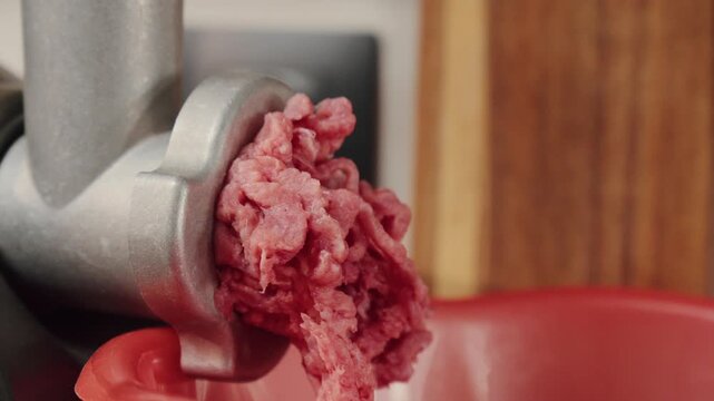 Meat grinding process. Closeup of raw meat exiting grinder into bowl. Handoperated grinder forces fresh minced meat into vibrant red container