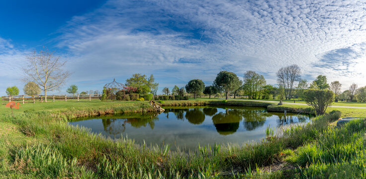 Charencey, France - 04 10 2026: Panoramic view of a pond, large trees, grass and a blue cloudy and sunny sky in a park in Le Perche region