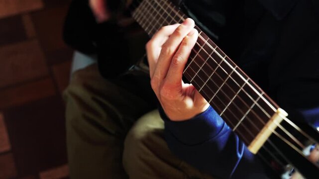 A person sits and plays guitar indoors. The hand is pressing strings