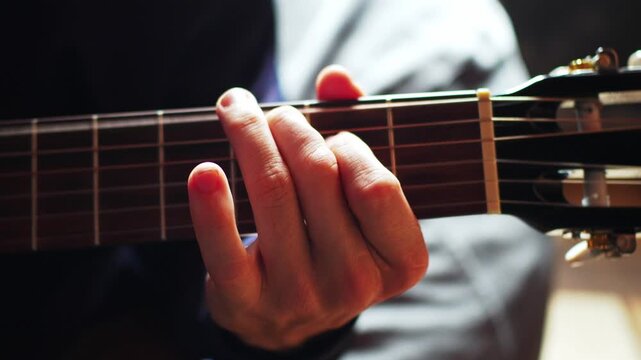 A person sits in a dimly lit room, focused on playing a guitar. The hand presses down on the strings