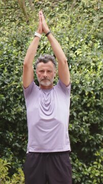 Vertical video: Starting arms up purple tee man lowering hands to chest in yard for yoga with watch
