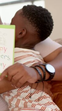 Vertical video: Son in striped tee holding green card, leaning into father at home for Fathers Day