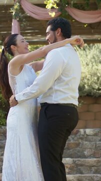 Vertical video: Smiling couple leaning into embrace on steps under pergola, in lace gown at wedding