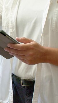 Vertical video: Framing adult in white tee adjusting, tapping folio tablet by window while reading