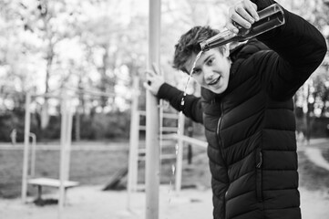 Teenage boy pours liquid from bottle in playground area. Troubled youth in black jacket shows...