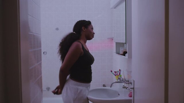 African American woman leaning toward bathroom mirror inspecting skin closely with concerned expression, focusing on facial imperfections, self image, and quiet dissatisfaction with appearance