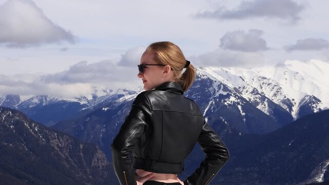 A girl on the top of a snow-capped mountain in a leather jacket and sunglasses.