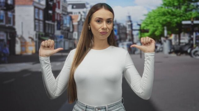 Woman with thumbs pointing to chest on a city street wearing white longsleeve top and jeans, long straight hair and gold studs, neutral smirk; confidence.